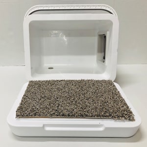 Puede incluir: Una caja de arena para gatos blanca y cerrada, con tapa con bisagras y puerta incorporada. La base de la caja tiene una alfombra rectangular, texturizada, marr&oacute;n y gris. La caja de arena est&aacute; dise&ntilde;ada para contener olores y brindar privacidad al gato.
