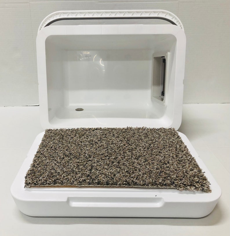 Puede incluir: Una caja de arena para gatos blanca y cerrada, con tapa con bisagras y puerta incorporada. La base de la caja tiene una alfombra rectangular, texturizada, marr&oacute;n y gris. La caja de arena est&aacute; dise&ntilde;ada para contener olores y brindar privacidad al gato.