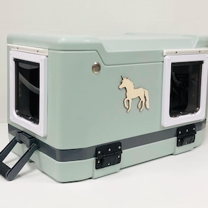 May include: A light green pet carrier with two windowed openings framed in white. A wooden unicorn is affixed to the side. The carrier has a dark gray handle and hinges, and a small circular vent.