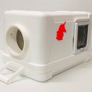 May include: A white, rectangular cat litter box with a circular opening and a small pet door. A red unicorn silhouette is affixed to the side. The box has a handle and is designed for pet use.