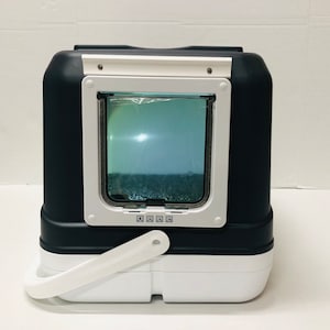 Puede incluir: Un arenero para gatos negro y blanco con una puerta rectangular transparente. La puerta est&aacute; enmarcada en blanco y tiene un peque&ntilde;o asa. El arenero tiene un asa blanca en la parte inferior y una parte superior blanca.