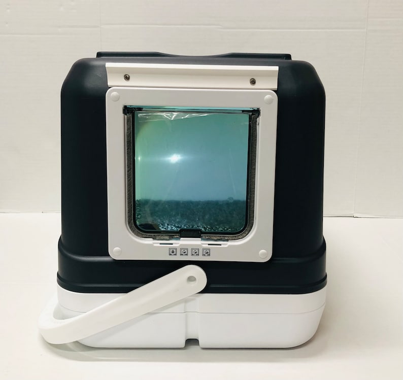 Puede incluir: Un arenero para gatos negro y blanco con una puerta rectangular transparente. La puerta est&aacute; enmarcada en blanco y tiene un peque&ntilde;o asa. El arenero tiene un asa blanca en la parte inferior y una parte superior blanca.