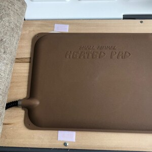 Puede incluir: Una almohadilla t&eacute;rmica marr&oacute;n para animales peque&ntilde;os con el texto "SMALL ANIMAL HEATED PAD" en relieve en la superficie. La almohadilla es rectangular con esquinas redondeadas y est&aacute; sujeta a una base de madera con tiras de velcro. Una esterilla beige enrollada est&aacute; al lado de la almohadilla.