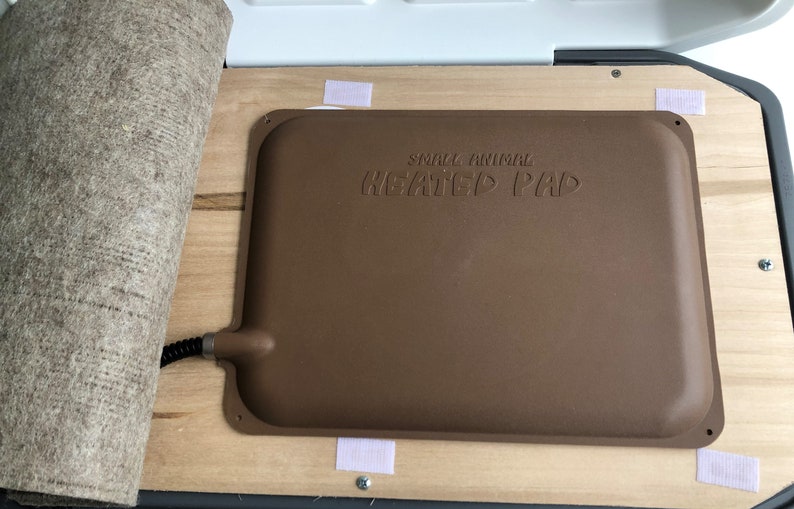 Puede incluir: Una almohadilla t&eacute;rmica marr&oacute;n para animales peque&ntilde;os con el texto "SMALL ANIMAL HEATED PAD" en relieve en la superficie. La almohadilla es rectangular con esquinas redondeadas y est&aacute; sujeta a una base de madera con tiras de velcro. Una esterilla beige enrollada est&aacute; al lado de la almohadilla.