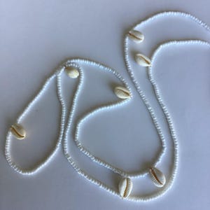以下が含まれることがあります： 白いビーズのネックレスに、カウリ貝が飾られています。ネックレスは2連になっていて、カウリ貝は全部で7つ付いています。