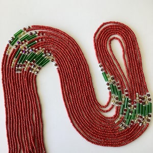 May include: A long, red beaded necklace with green and gold accents. The necklace is made of multiple strands of beads and is arranged in a decorative pattern.