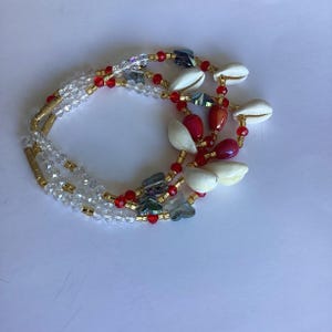 May include: A multi-strand necklace featuring clear crystal beads, red beads, gold-colored tubes, and white seashells. The necklace has a layered design with a mix of textures and colors, creating a unique accessory.