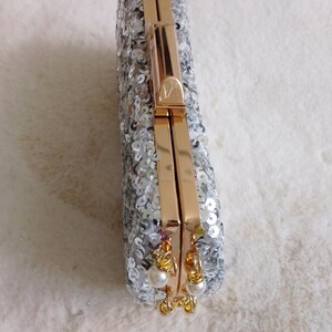 May include: A rectangular silver sequined clutch with gold-colored hardware. The clutch has a clasp closure and small pearl accents hanging from the bottom. The bag is on a white, fluffy surface.