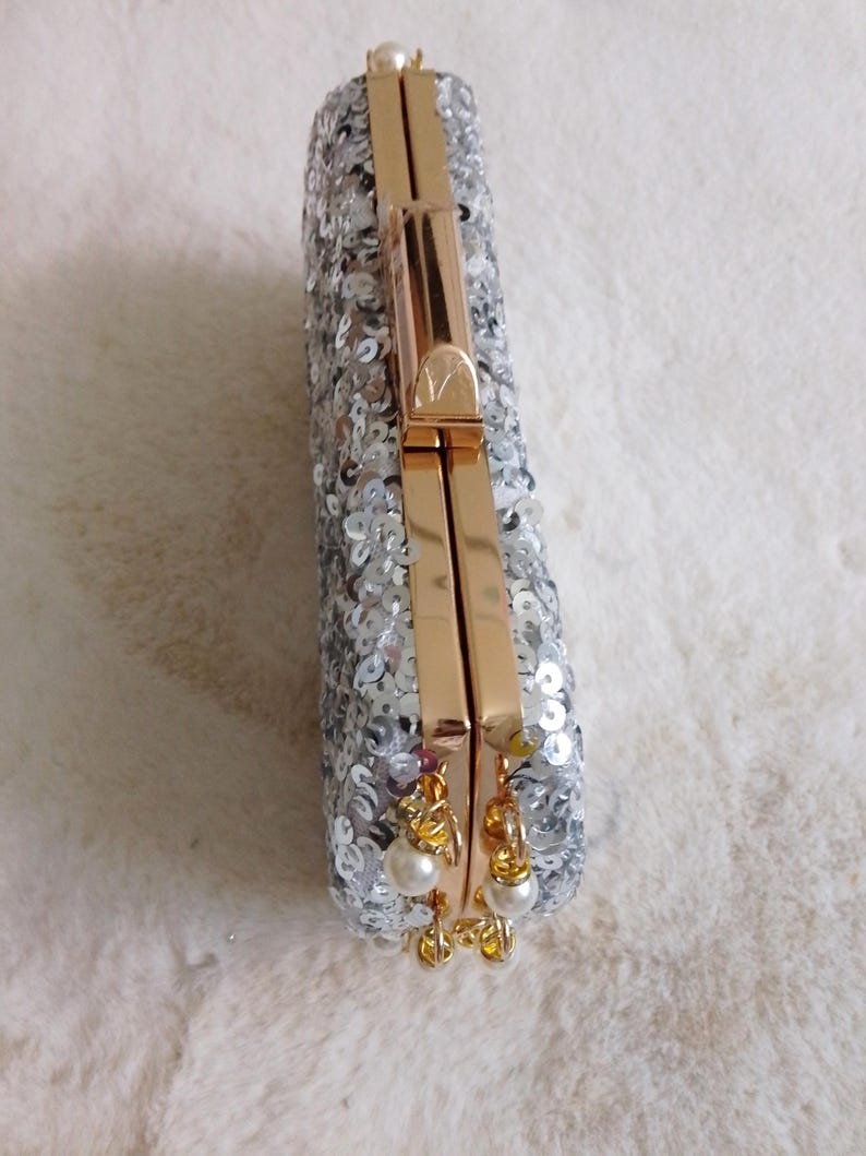 May include: A rectangular silver sequined clutch with gold-colored hardware. The clutch has a clasp closure and small pearl accents hanging from the bottom. The bag is on a white, fluffy surface.