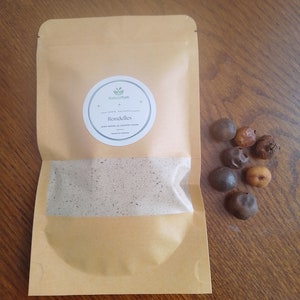 May include: A brown paper bag with a white circle label that says "NaturaYum 100% Natural Rondelles Also known as Country Onion Product of Cambodia" and a small green leaf icon. There are several brown and tan round objects scattered on a wooden surface next to the bag.