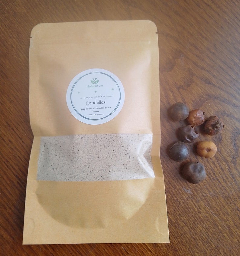 May include: A brown paper bag with a white circle label that says "NaturaYum 100% Natural Rondelles Also known as Country Onion Product of Cambodia" and a small green leaf icon. There are several brown and tan round objects scattered on a wooden surface next to the bag.