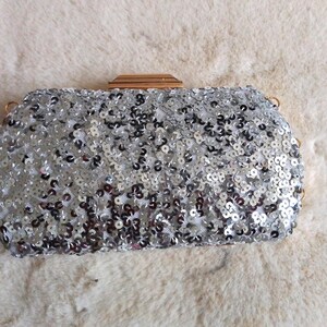 May include: Silver sequin clutch with a rectangular shape and a gold-toned clasp. The bag is covered in small, reflective sequins, creating a shimmering effect. The clutch has a chain strap and is set against a soft, white background.