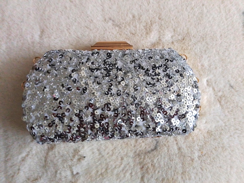 May include: Silver sequin clutch with a rectangular shape and a gold-toned clasp. The bag is covered in small, reflective sequins, creating a shimmering effect. The clutch has a chain strap and is set against a soft, white background.