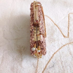 May include: Rose gold sequined clutch with a gold chain strap. The rectangular evening bag features a gold clasp and is adorned with small white pearl accents. The clutch is designed for formal events.