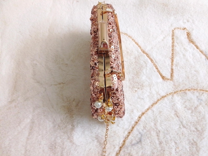 May include: Rose gold sequined clutch with a gold chain strap. The rectangular evening bag features a gold clasp and is adorned with small white pearl accents. The clutch is designed for formal events.