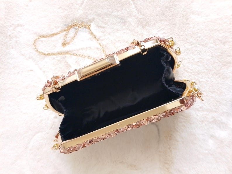 May include: Open rose gold clutch with a gold chain strap. The clutch is covered in sequins and has a gold clasp. The interior is black. A stylish accessory for special occasions.