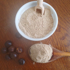 May include: A white bowl filled with light brown powder, a wooden spoon with a handle sticking out of the bowl.  A small pile of dark brown seeds are scattered on a wooden surface.