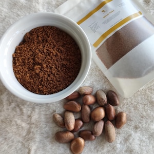 May include: A white bowl filled with brown powder, a white bag with the text "Whole P&b" and a pile of brown and tan seeds on a white fluffy surface.