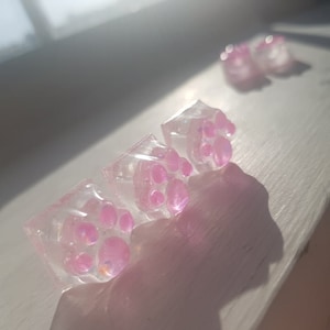 Może przedstawiać: Cztery przezroczyste klawisze z żywicy z różowymi odciskami łapek. Klawisze są przezroczyste i mają błyszczące wykończenie.