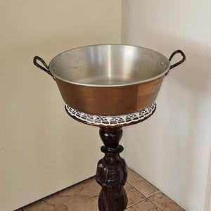 May include: A copper and aluminum bowl with two handles sits on a wooden stand with a carved figure. The stand has a white crocheted doily on top.