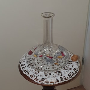 May include: A clear glass decanter with a decorative design sits on a wooden table with a carved figurine of a person. The table has a white crocheted doily on top.