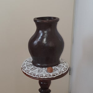 Op de afbeelding: Een donkerbruine keramische vaas staat op een houten sokkeltafel met een wit gehaakt tafellaken. De tafel heeft een gegraveerde houten basis in de vorm van een persoon.