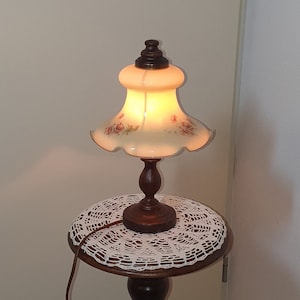 Puede incluir: Una lámpara de mesa vintage con base de madera oscura y pantalla con estampado floral. La lámpara tiene una mesa redonda con un tapete de encaje blanco. La lámpara mide aproximadamente 140 cm de altura.
