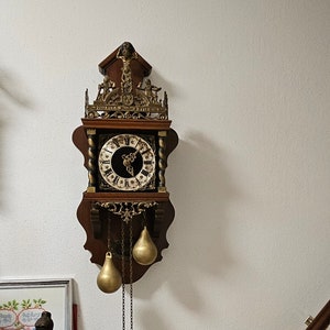 Puede incluir: Un reloj de pared antiguo de madera con adornos dorados y una esfera negra. El reloj tiene dos campanas doradas colgando de cadenas.