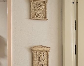 May include: Two rectangular wall plaques with classical relief designs. The plaques are beige and feature detailed figures within a framed architectural setting. The top plaque depicts a figure pulling a bow, while the bottom one shows two figures embracing.