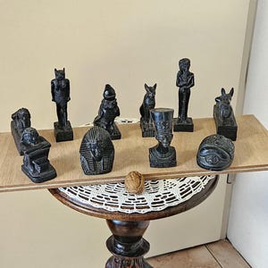 Op de afbeelding: Een verzameling kleine, zwarte Egyptische beeldjes tentoongesteld op een houten tafel. De beeldjes beelden verschillende farao's en dieren uit. De tafel heeft een decoratieve, gesneden houten voet en een wit kanten kleedje.