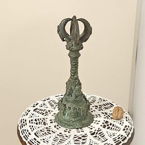 Op de afbeelding: Een antiek bronzen decoratief object met een gedetailleerd ontwerp, mogelijk een religieus artefact, staat op een kleine houten tafel met een wit kanten kleedje. Een walnoot ligt op de tafel. De tafel heeft een gebeeldhouwde figuur als basis.