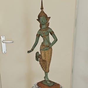 May include: A bronze-colored statue of a dancing figure with ornate headdress and clothing, mounted on a wooden base. The statue is displayed on a small, dark wooden table with a lace doily. The figure has a green patina.