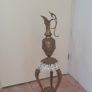 Op de afbeelding: Een decoratieve standaard van goudkleurig metaal met een ronde bovenkant en drie poten. De standaard heeft een wit kantkleedje op de bovenkant en een gouden metalen kan met ingewikkelde details.