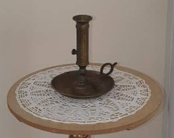 May include: A gold metal stand with a round top and three legs. A brass candlestick with a white doily sits on the top.