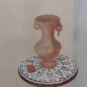 May include: A wooden pedestal table with a carved figure standing on the column. The table has a white crocheted doily on top with a terracotta vase.
