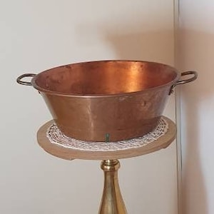 May include: A copper bowl with two handles sits on a gold metal stand with a round top and three legs. The stand is on a tiled floor.