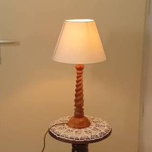 May include: A wooden table lamp with a white lampshade sits on a wooden pedestal table with a white lace tablecloth. The lamp is turned on and the light is shining.