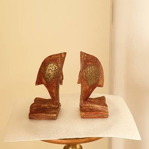以下が含まれることがあります： 彫刻が施され、金色のアクセントが付けられた木製のブックエンドのペア。ブックエンドは金色の台座の上に置かれています。