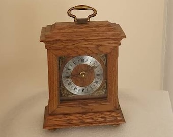 May include: A wooden clock with a brass handle on top and a white face with black Roman numerals. The clock is sitting on a white pedestal with a gold base.
