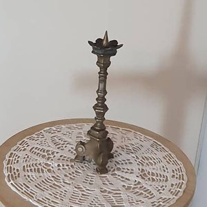 May include: A gold metal candlestick stand with a round wooden top and a white crocheted doily. The candlestick is made of brass and has a decorative design.