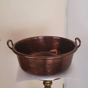 May include: A copper basin with two handles sits on a gold metal stand. The basin is round and has a slightly hammered texture. The stand has a decorative base and a tall, slender column.