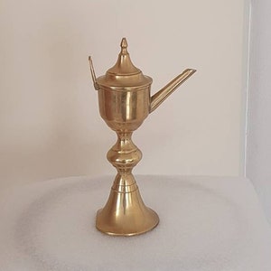 Op de afbeelding: Een gouden metalen olielamp met een decoratief ontwerp. De lamp heeft een ronde basis en een tuit om olie in te gieten.