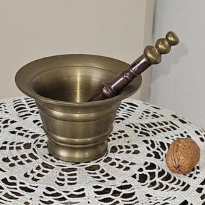 May include: A vintage side table made of dark wood, with a carved figure base. It has a brass mortar and pestle, a white crocheted doily, and a walnut. The table is a decorative item.