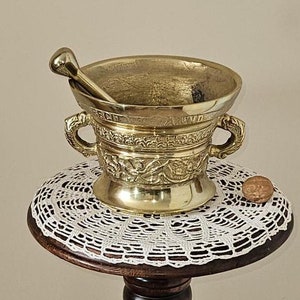May include: A gold mortar and pestle set on a wooden stand with a carved figure. The stand has a white lace doily on top.