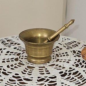 May include: A small, antique brass mortar and pestle sit on a round, dark wooden table. The table is covered with a white, crocheted doily. A walnut rests on the doily. The table has a carved wooden base.