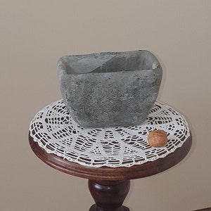 Op de afbeelding: Een houten voetstuktafel met een gegraveerd figuur van een man met een hoed. De tafel heeft een ronde bovenkant met een wit gehaakt tafellaken en een grijze rechthoekige plantenbak erop.