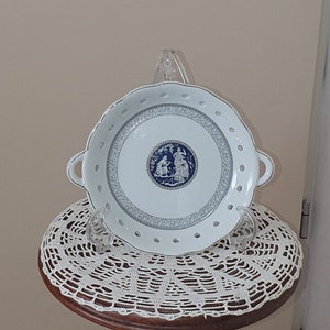 May include: A white porcelain plate with a blue and white floral design sits on a white crocheted doily on a wooden pedestal stand. The stand is carved with a figure of a person.