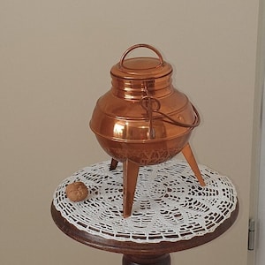 May include: A copper pot with a lid and three legs sits on a wooden table with a white doily. The table is supported by a carved wooden figure.
