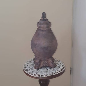 May include: A brown ceramic vase with a decorative top sits on a wooden pedestal table with a white crocheted doily. The table has a carved wooden base in the shape of a person.