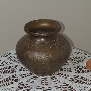 May include: A round, ornate, brass vase sits on a white crocheted doily on a wooden table with a carved figure of a person standing on the base.